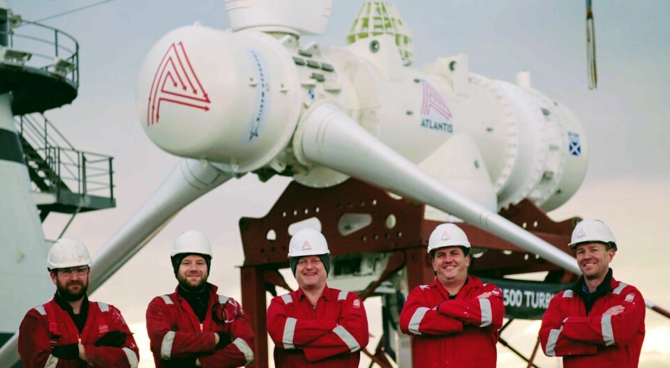Fem män med hjälmar poserar framför en stor turbin med tre rotorblad.