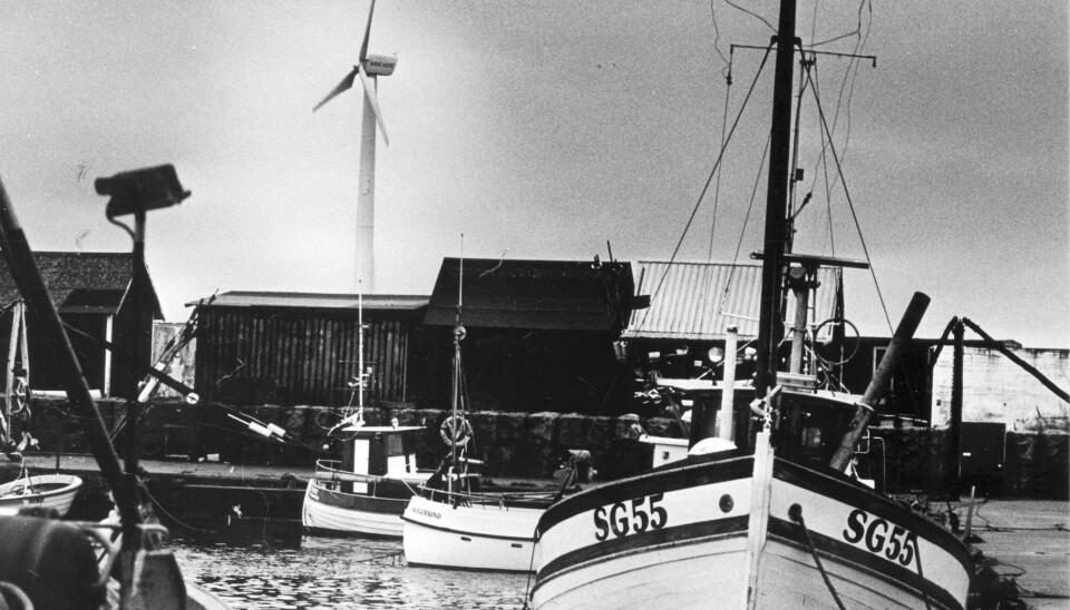 En svartvit bild där vindkraftverket syns bakom en äldre träskuta och byggnader i en hamn.