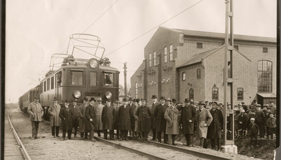 Ett elektriskt lok av den äldre stilen med träpanel längs fram. Framför tågsättet står en mängd finklädda herrar uppställda.
