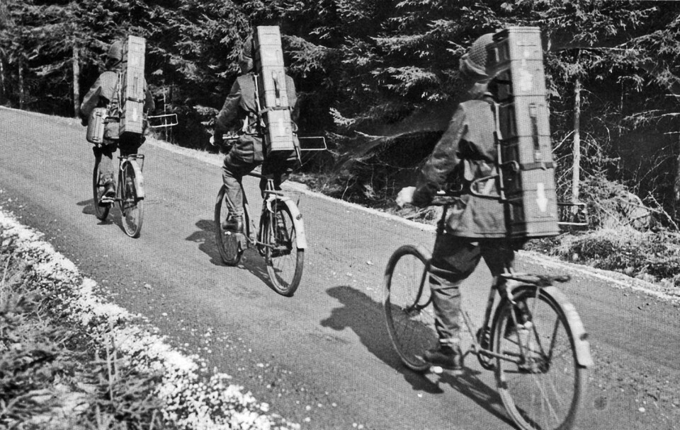 Tre soldater i uniform som cyklar med varsin Robot 54 i en rektangulär låda på ryggen. De cyklar på en mindre grusväg.
