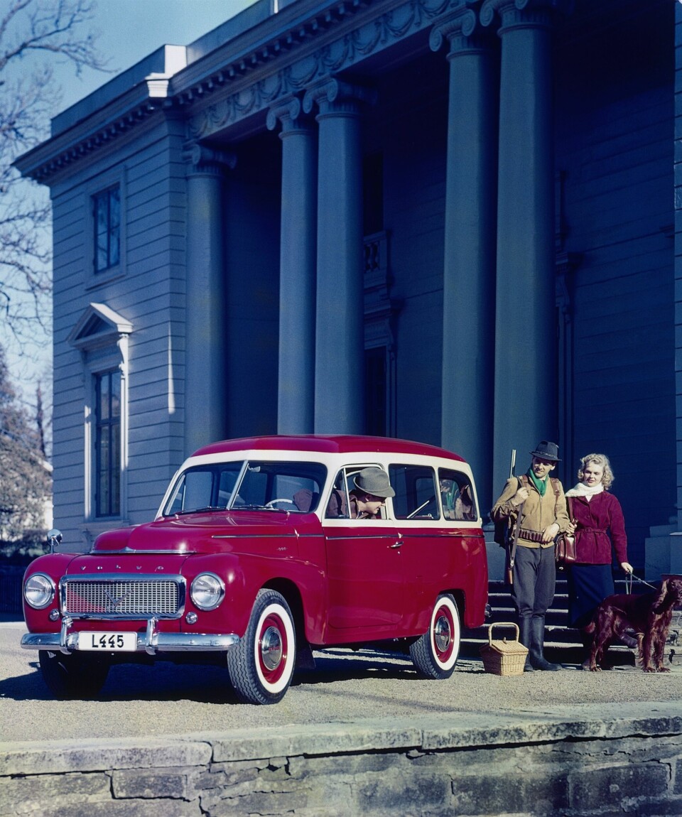 En vinröd Volvo Duett med vita fönsterramar står redo för avfärd utanför ett hus. En man med ett gevär över axeln och en kvinna som håller en hund står bredvid