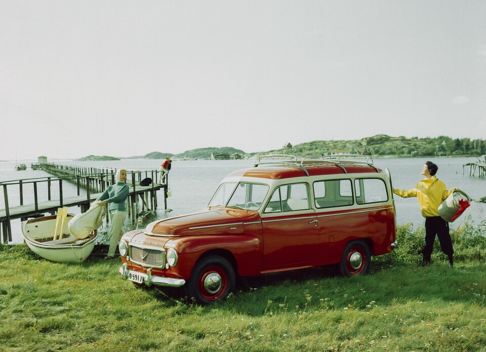 En röd Volvo Duett parkerad på en gräsmatta intill en brygga. Två kvinnor i fritidskläder packar ur campingutrustning.