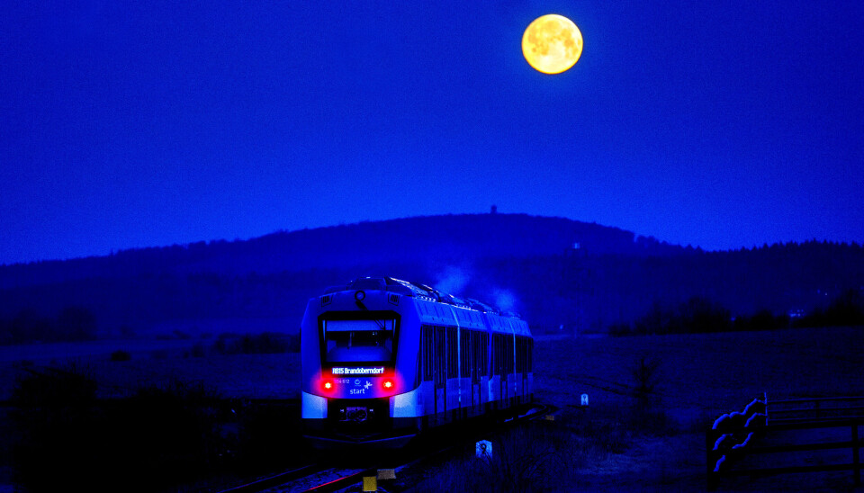 Ett vätgaståg lämnar stationen i Wehrheim nära Frankfurt, Tyskland. Tåg med röda lampor framför en blå natthimmel som visar en gul fullmåne.