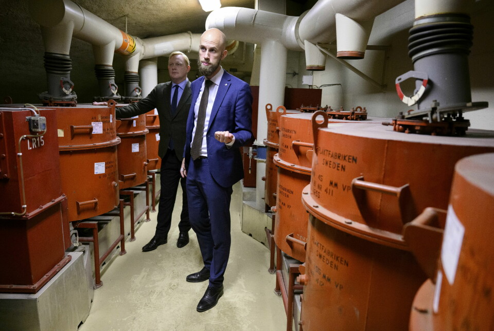 Man without hair in a blue suit stands in a basement room surrounded by large red containers.