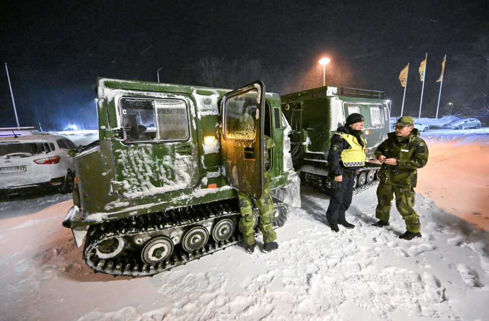 En militärt grönmålad bandvagn i snöyran vid motorvägen i Skåne med två män i kamouflagekläder och en man i gul varselväst som står och pratar utanför.