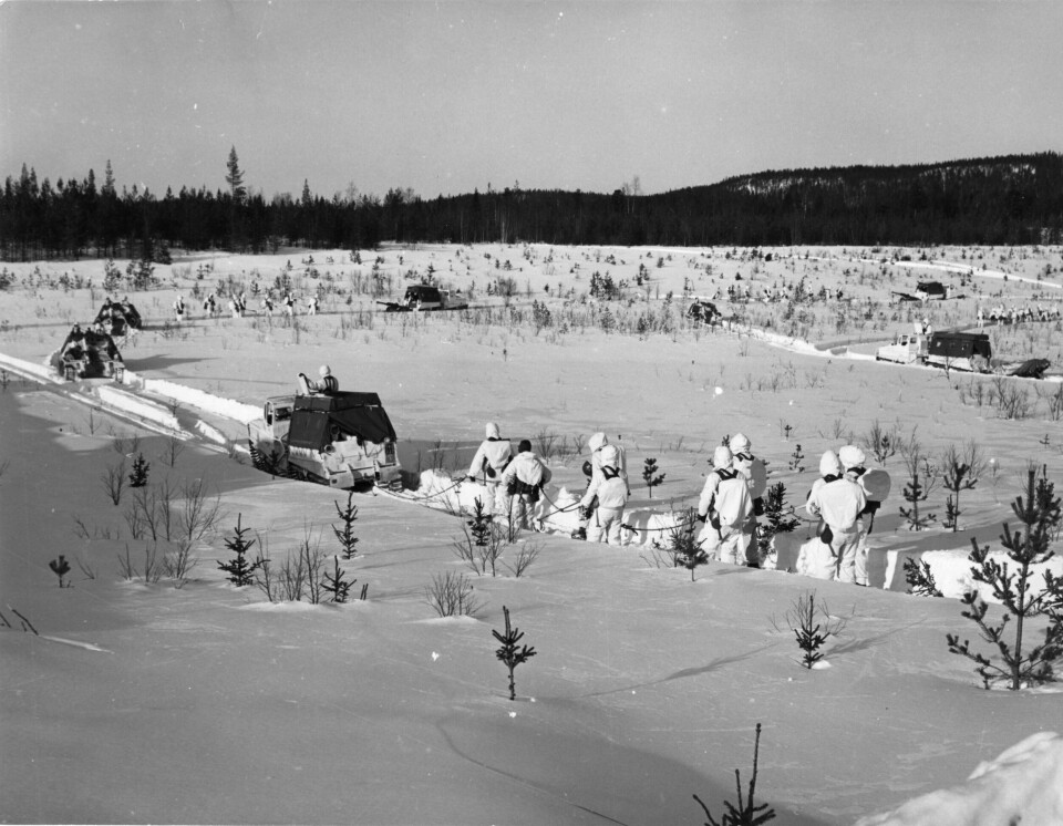 Soldater i vita kamouflagedräkter tolkar på skidor i djup snö, i bakgrunden syns en höjd med granskog.