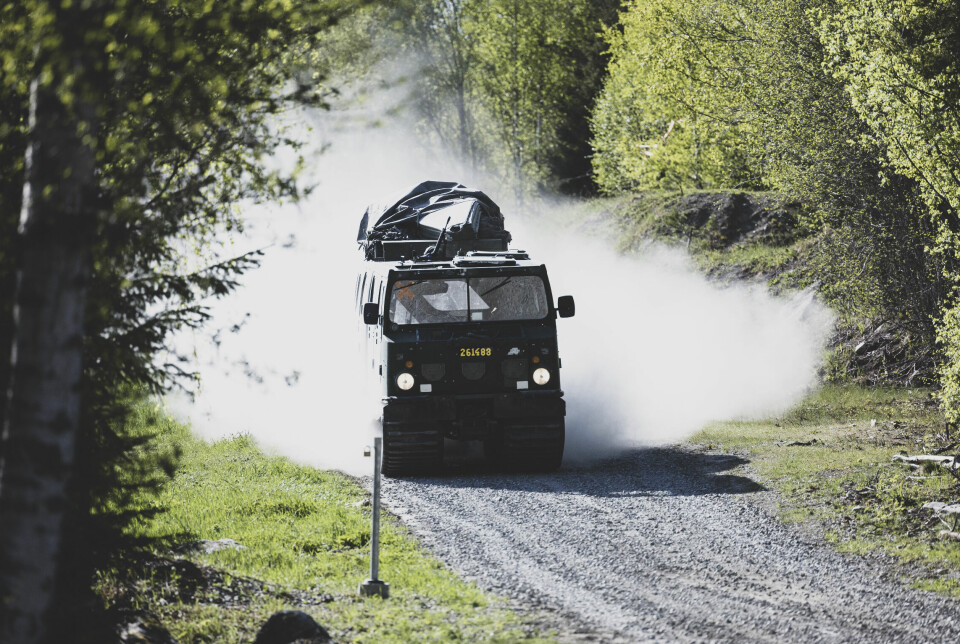 En militärt målad bandvagn kör i full fart på en grusväg, med ett moln av damm bakom sig.