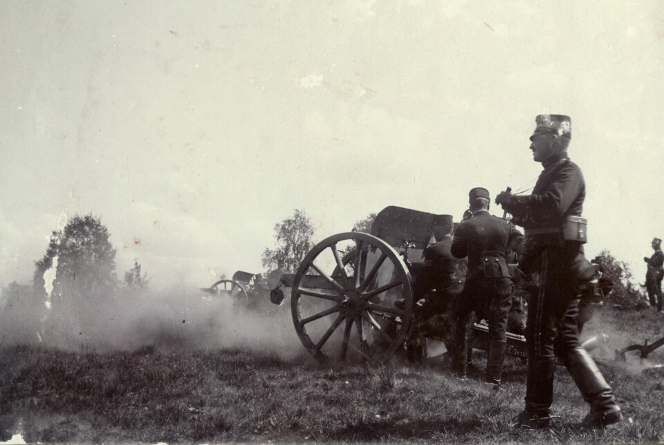 Krutrök sprids runt en kanon direkt efter en avfyrning, bilden är svartvit och det står en soldat i militära kläder i förgrunden