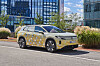 Kamouflerad Volkswagen elbil parkerad på en gata framför moderna kontorskomplex av glas.